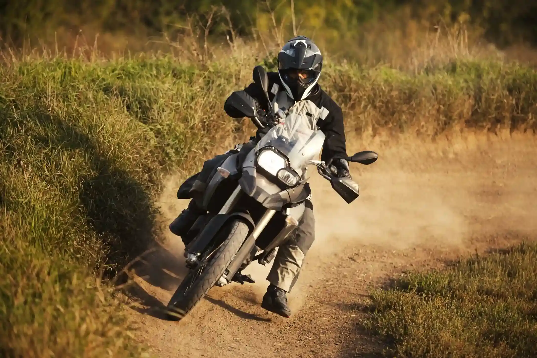 A rider on an adventure motorcycle leans through a dirt-track corner, kicking up a dust cloud in tall dry grass.