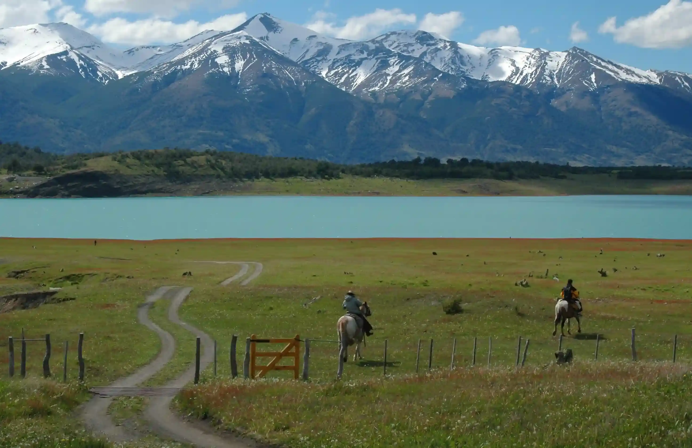 Two gauchos on horseback crossing a wooden gate at a Patagonian estancia, with a turquoise glacier-fed lake and snow-capped Andes in the background.