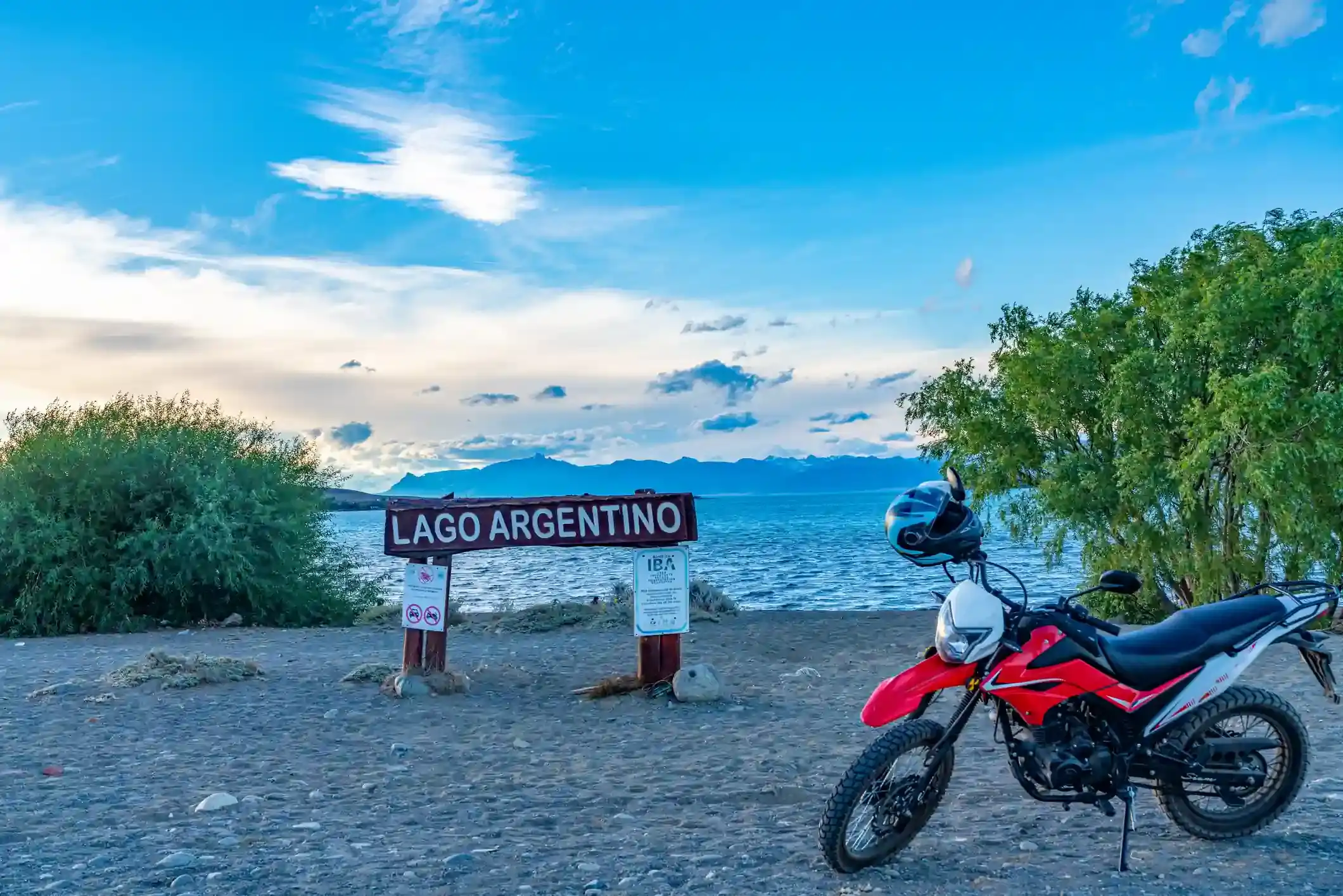 A motorcycle parked beside the Lago Argentino sign in El Calafate, with the Patagonian lake and low mountains behind at dusk.