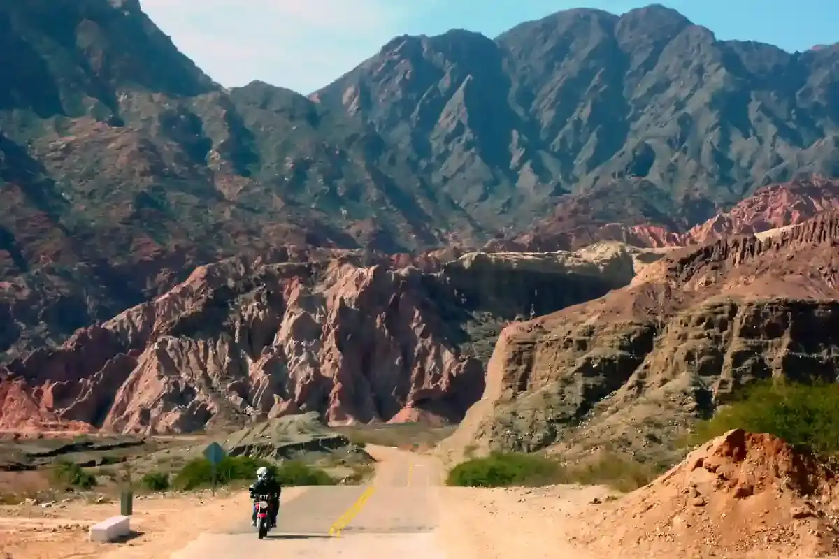 A motorcycle rider on a paved road approaching the red rock formations of the Quebrada de las Conchas near Cafayate, Argentina.