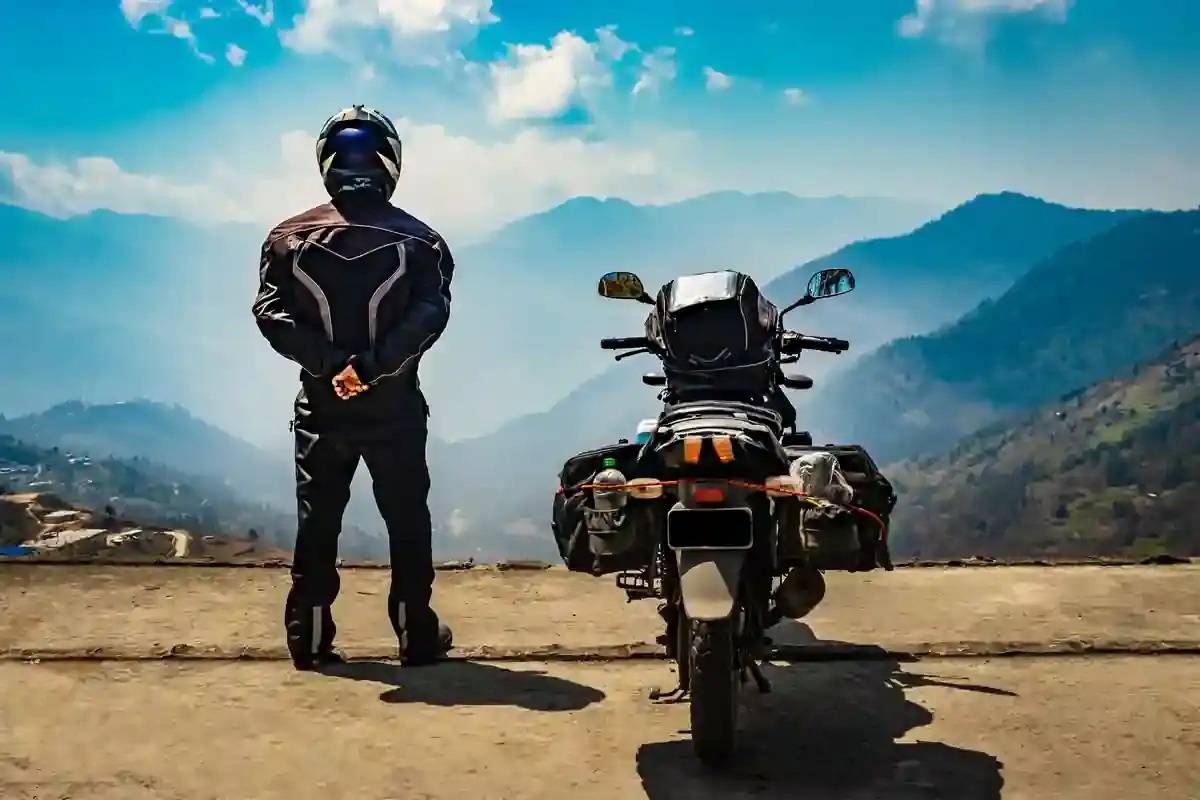 A rider in full gear stands beside a loaded adventure motorcycle at an overlook, facing layered mountain ridges.